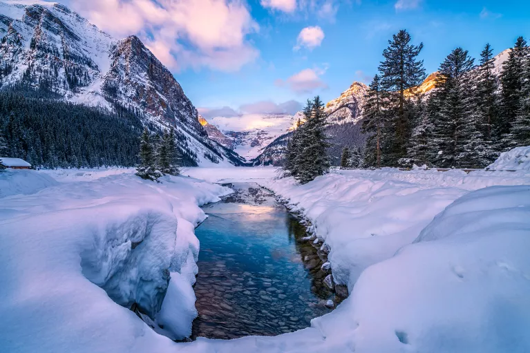 Wide shot of snowy valley, mountains, trees around.