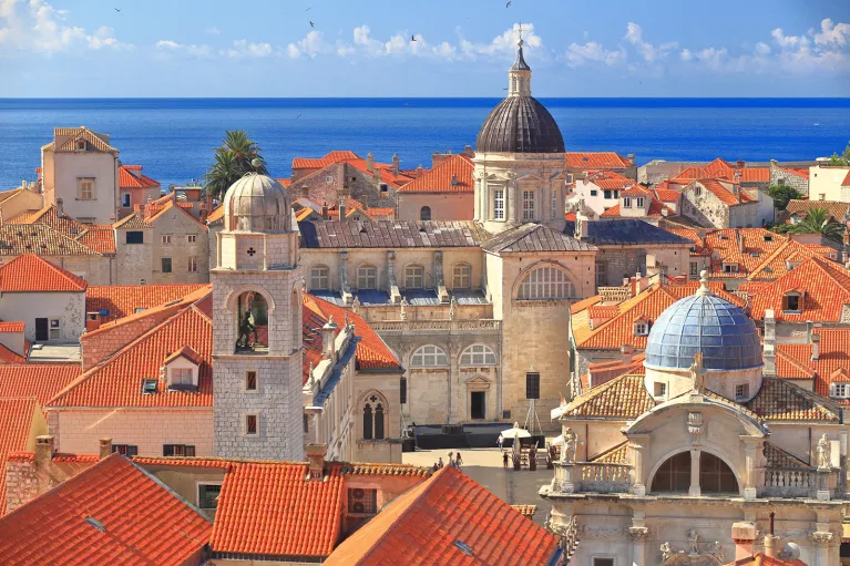 Wide shot of Dubrovnik coastline, blue ocean, white and tan houses.
