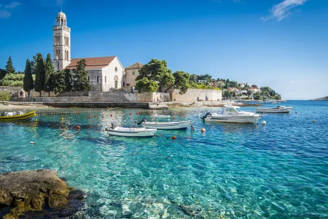 Wide shot of Hvar Island, white stone buildings, blue water, boats.