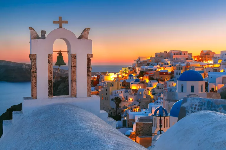 Sunset shot of Santorini, city lit up, church bell in foreground.