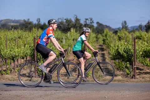 Two guests on e-bikes riding past vineyard.