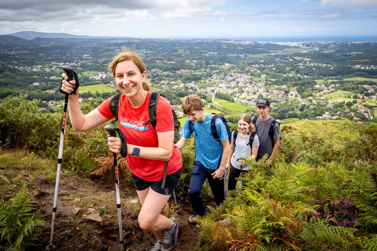 Four guests walking up hillside trail, French village, coast in distance. 