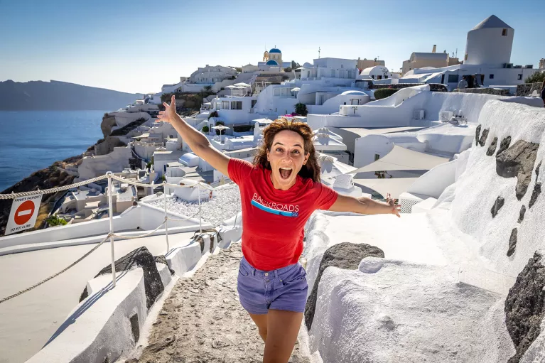 Guest emoting to camera, white walled coastal buildings behind her.