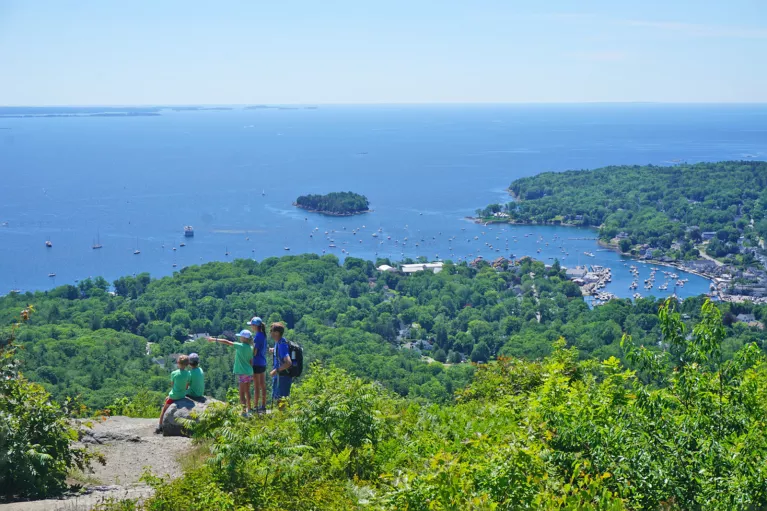 Wide shot of a coastal vista, sailboats visible in water, group of young guests in foreground.