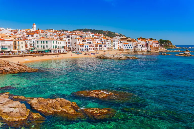 Shot of Costa Brava coastline, blue water, tan and white buildings.