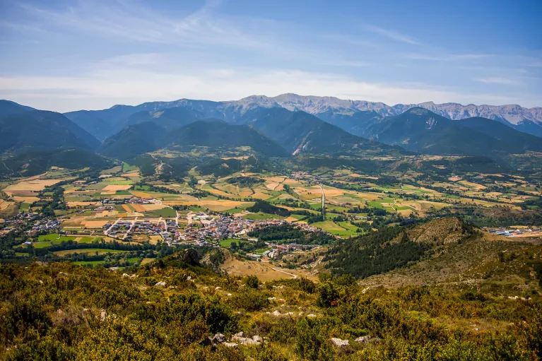 Bird's eye shot of mountainous valley, town, golden fields below.
