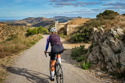 Guest cycling down gravel road, towards stone ruins, golden hills. 