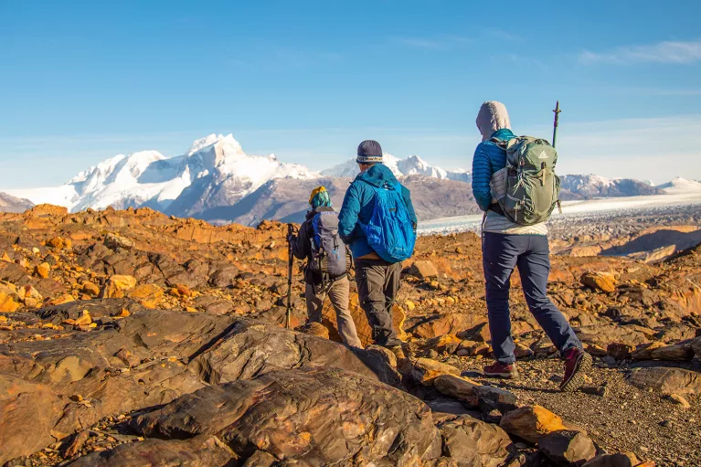 Three guests walking over orange rocks, snowy peaks in distance.