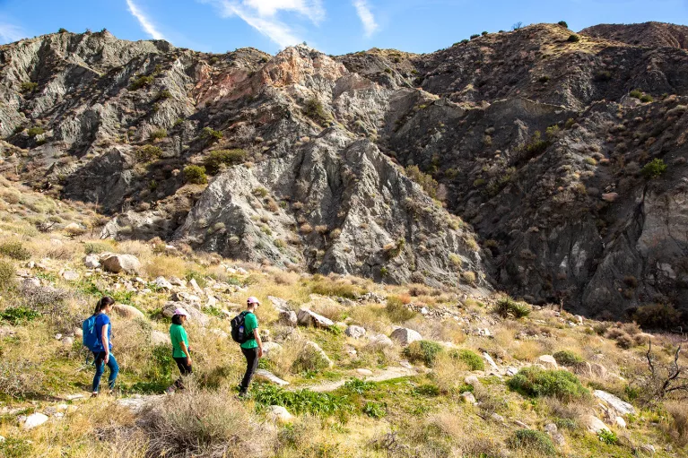 Group of three hiking in desert, craggy hill in background.
