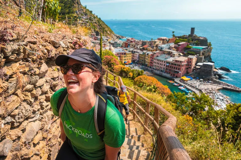 Two guests hiking up rocky trail, colorful coastal town in background.