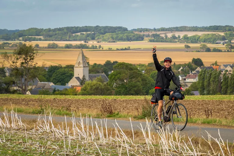 Man biking on an asphalt road in a grass valley, while he's waving his hand in the air