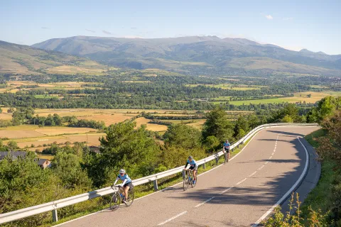 Group of 3 people riding bikes on an asphalt road, with valleys of trees in the distance