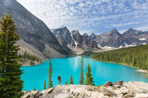 Man standing on a boulder, looking out to a blue lake and tall trees