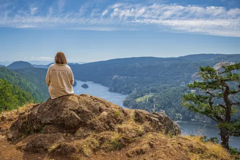 Woman sitting on a large boulder on top of a hill, looking down at the water