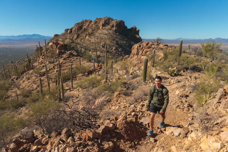 Man ascending a rocky, dirt trail with canyons and cacti behind him