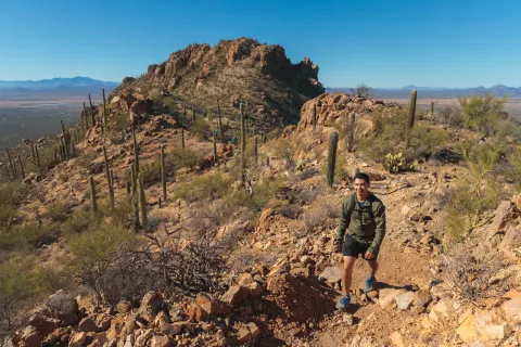 Man ascending a rocky, dirt trail with canyons and cacti behind him