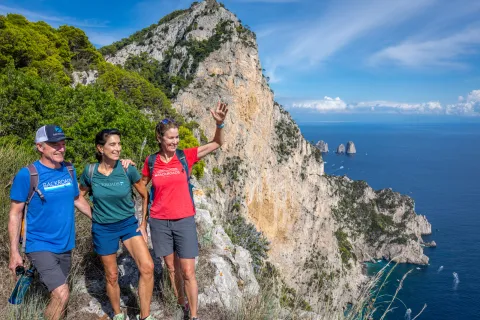 Two women and a man smiling and waving, while standing on a cliff next to the ocean