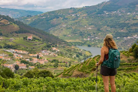 Woman wearing a backpack, standing on top of a hill looking down at a small town surrounded by trees