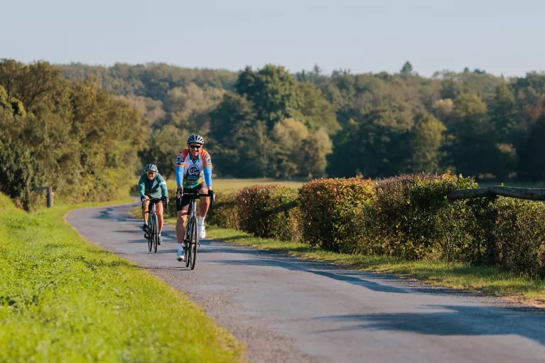 Two people smiling while riding their bikes on a road surrounded by bushes and trees