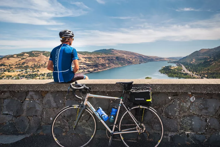 Man sitting on a stone ledge looking out to a lake, with a bike leaning on the ledge