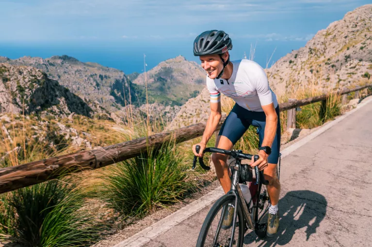 Man smiling while riding a bike on a road, with large cliffs in the distance
