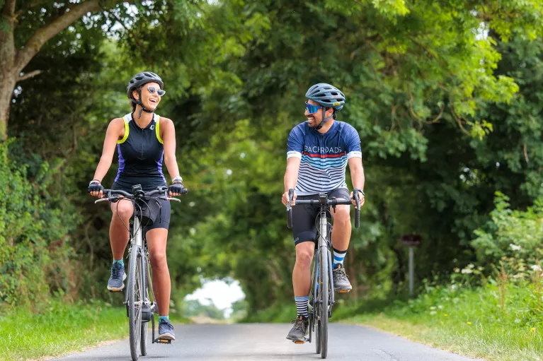 Man and woman smiling while riding their bikes on a road, with large trees surrounding