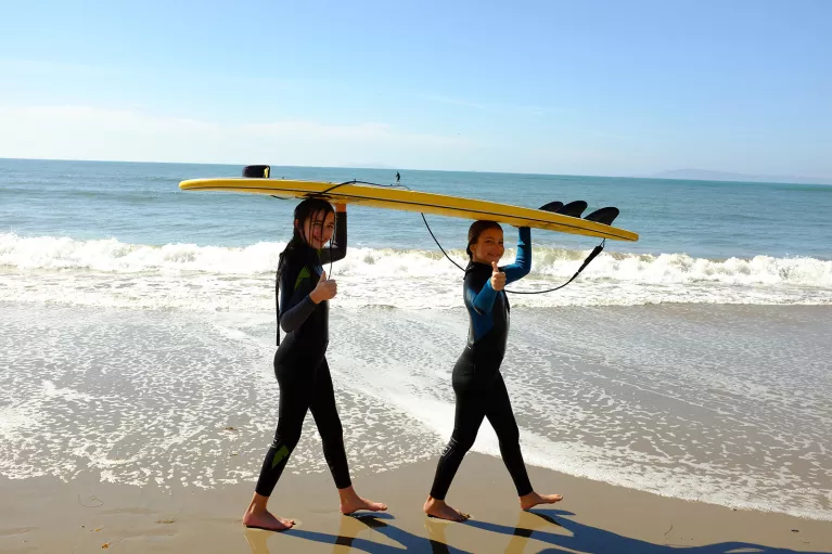 Two girls carrying a yellow surfboard over their head