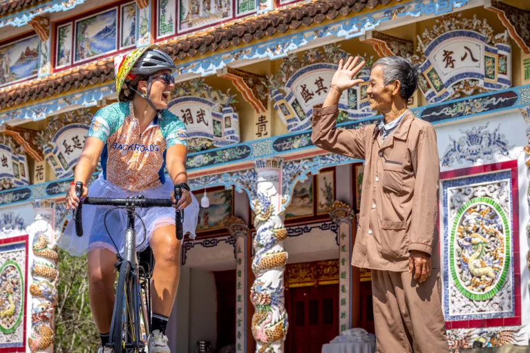 Woman riding a bike in front of a large temple, with a man smiling and waving at her
