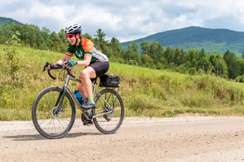 Woman riding a bike on a dirt road, with a grassy hill in the background