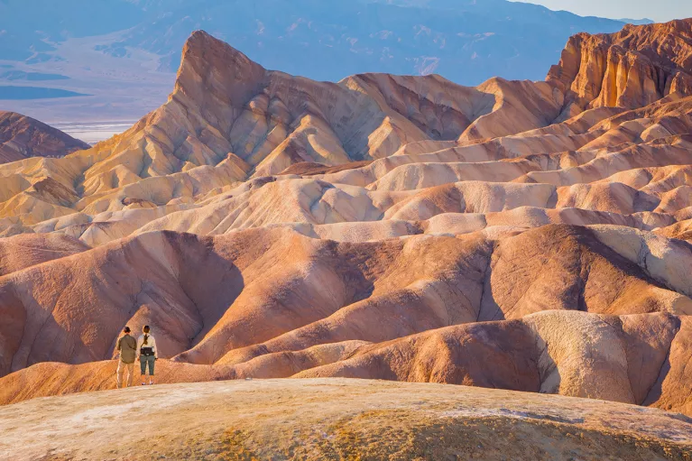 Two people standing on top of a large, orange canyon looking at larger canyons