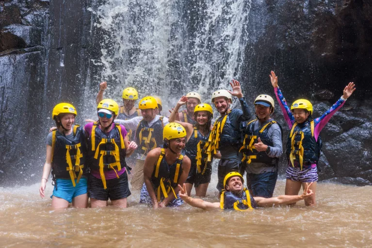 Group of people smiling while wearing helmets and life vests under a waterfall
