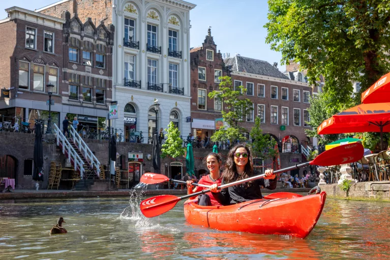 Two women paddling in a red kayak with a European town in the background