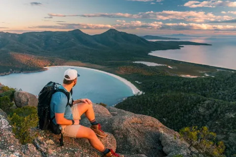 Man sitting on a cliff looking out to a valley of trees and a lake