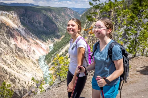 Two girls using hiking poles, walking on a dirt trail and large mountains in the distance