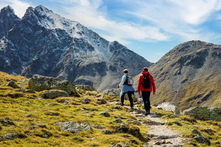 Man and woman ascending a rocky, dirt trail with large mountains ahead