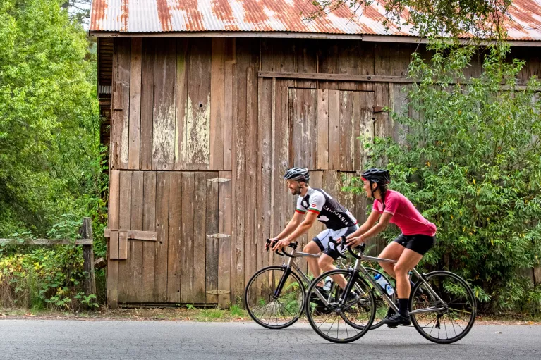 Man and woman wearing biking gear, riding bikes in front of a wooden barn on a road