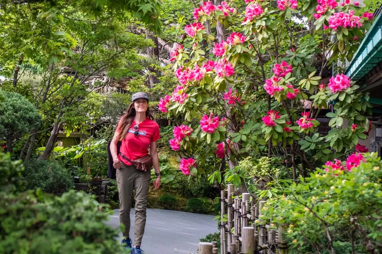 Woman smiling while walking through an outdoor garden full of plants and pink flowers