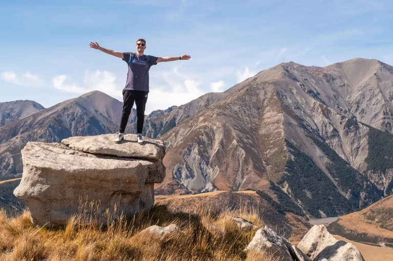 Man standing on top of a cliff with his arms wide open, with large mountains in the background