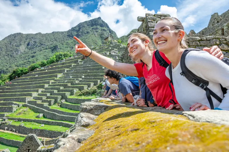Two women smiling and pointing towards the sky, with large stone ruins in the background