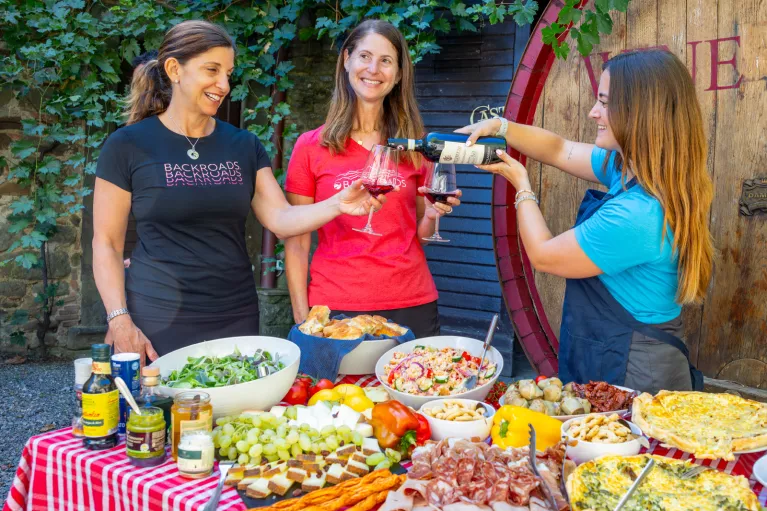 Three women behind a table full of food, pouring wine into glasses