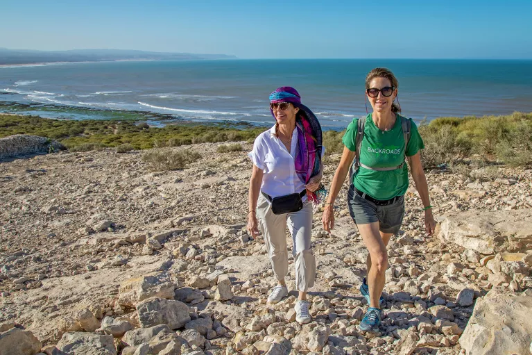 Two women smiling while walking on a gravel trail with the beach in the background