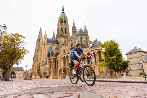 Cyclist rides across a cobblestone square in front of a Gothic cathedral featuring soaring spires, stained-glass windows, and ornate stone architecture in a historic French town.