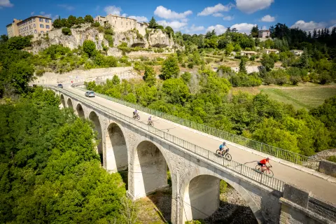 An aerial view of several cyclists crossing a large stone arch bridge above a wooded valley. A historic hilltop village with stone buildings sits on rocky cliffs in the background under a blue sky with scattered clouds.