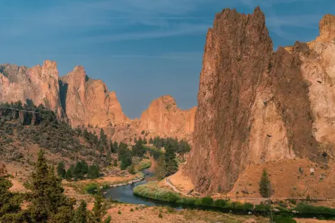 View of Smith Rock State Park featuring towering rock spires and cliffs rising above the Crooked River, which winds through a high desert canyon dotted with juniper trees.