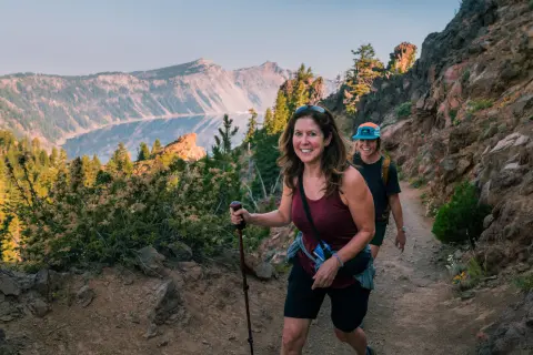 Two hikers walk along a rocky mountain trail overlooking a deep blue crater lake surrounded by rugged cliffs and evergreen trees. The scenic landscape is lit by soft natural light.