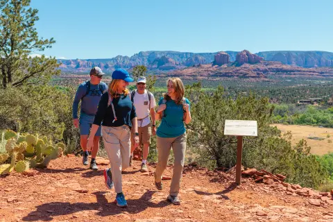 Group of hikers walking along a red dirt trail overlooking Sedona’s red rock formations and desert landscape under a clear blue sky.