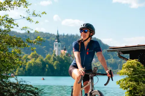 Cyclist pauses beside a turquoise lake with a church-topped island visible in the background. Forested hills and calm waters frame the scenic setting beneath a bright blue sky.