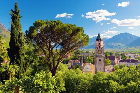 View of Merano, Italy, featuring a historic clock tower rising above lush gardens and trees. Mountain peaks and a broad alpine valley stretch into the distance beneath a bright blue sky dotted with white clouds.