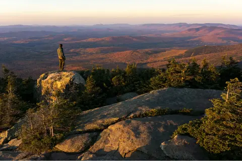 A hiker stands on a rock overlooking the sunlit mountain range in Vermont.