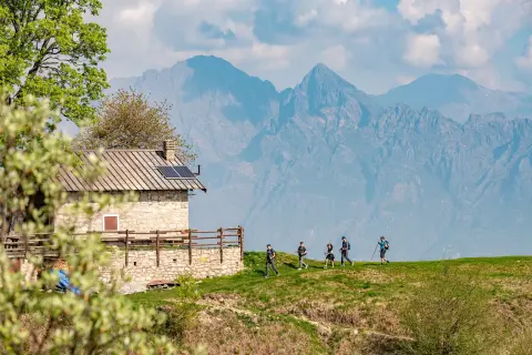 Hikers walk towards a stone home with a mountain range in the background.